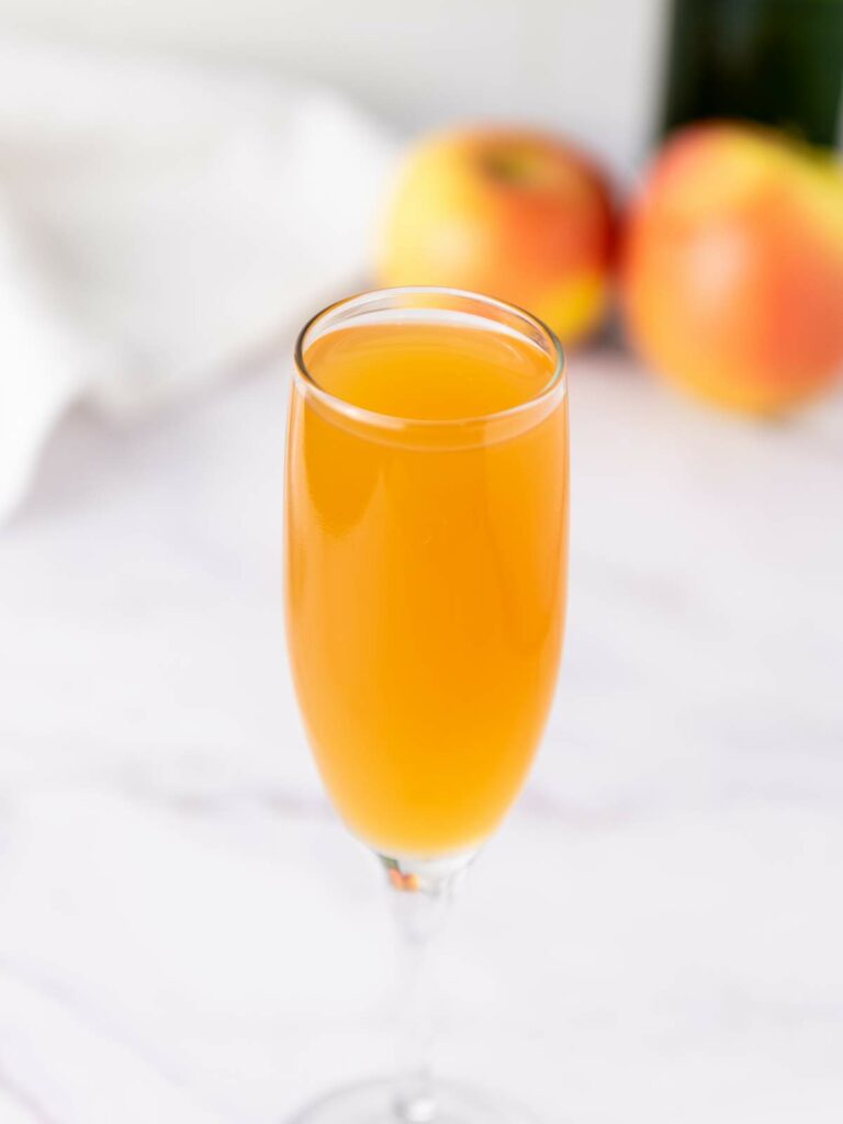 A glass of orange-colored drink in a champagne flute, with blurred apples and a white cloth in the background.