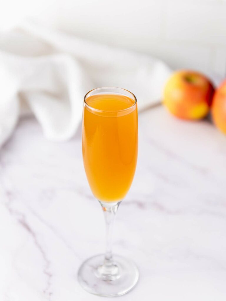 A champagne flute filled with cocktail sits on a marble surface, with two apples and a white cloth in the background.