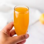 A hand holding a champagne flute filled with orange-colored juice against a light, blurred background.