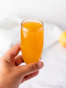 A hand holding a champagne flute filled with orange-colored juice against a light, blurred background.