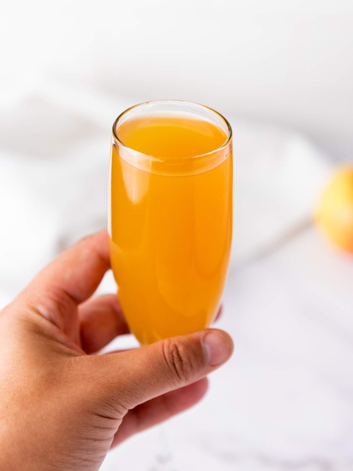 A hand holding a champagne flute filled with orange-colored juice against a light, blurred background.