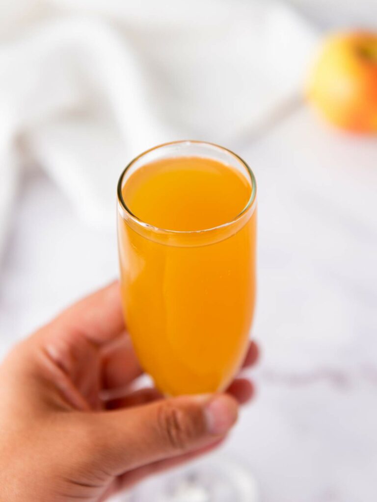 A hand holding a glass of orange-colored juice, with a blurred apple and white cloth in the background.