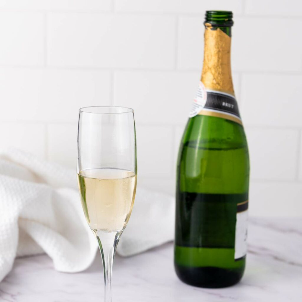A glass of champagne sits on a marble surface next to an open green champagne bottle.