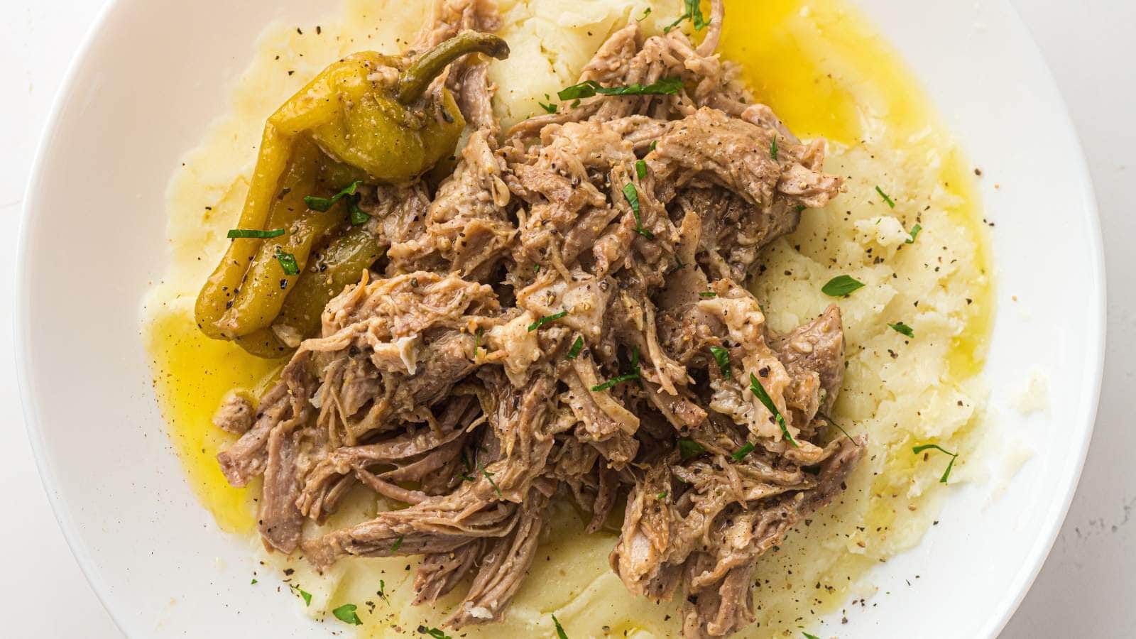 Shredded cooked meat served over mashed potatoes with a whole pepper. garnished with chopped herbs on a white plate.