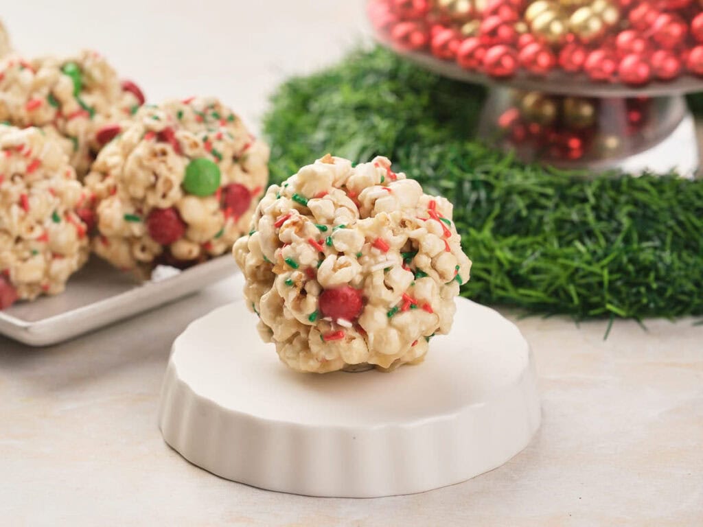 A popcorn ball with red and green chocolates and sprinkles; more popcorn balls and festive decorations are in the background.