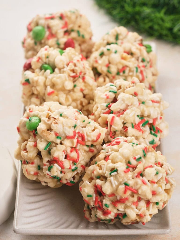 Six popcorn balls with white, red and green sprinkles and m&m's chocolate are arranged on a rectangular white plate.