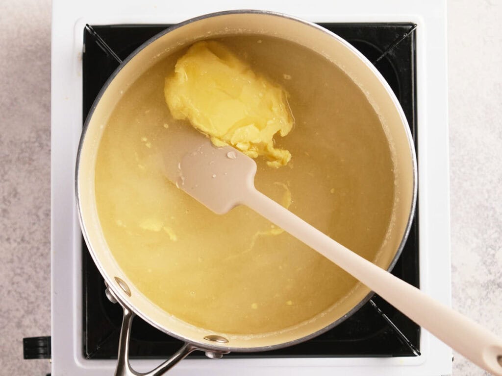 A saucepan filled with yellow liquid and a large chunk of butter being stirred with a spatula on a stovetop.