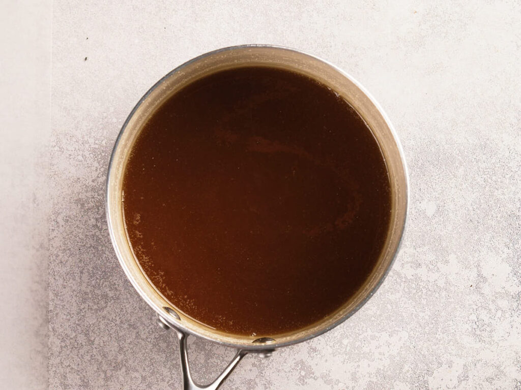 A stainless steel saucepan filled with dark brown liquid sits on a light-colored textured surface.
