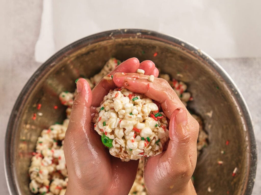 Hands shaping a popcorn ball with colorful sprinkles over a metal mixing bowl containing more popcorn mixture.
