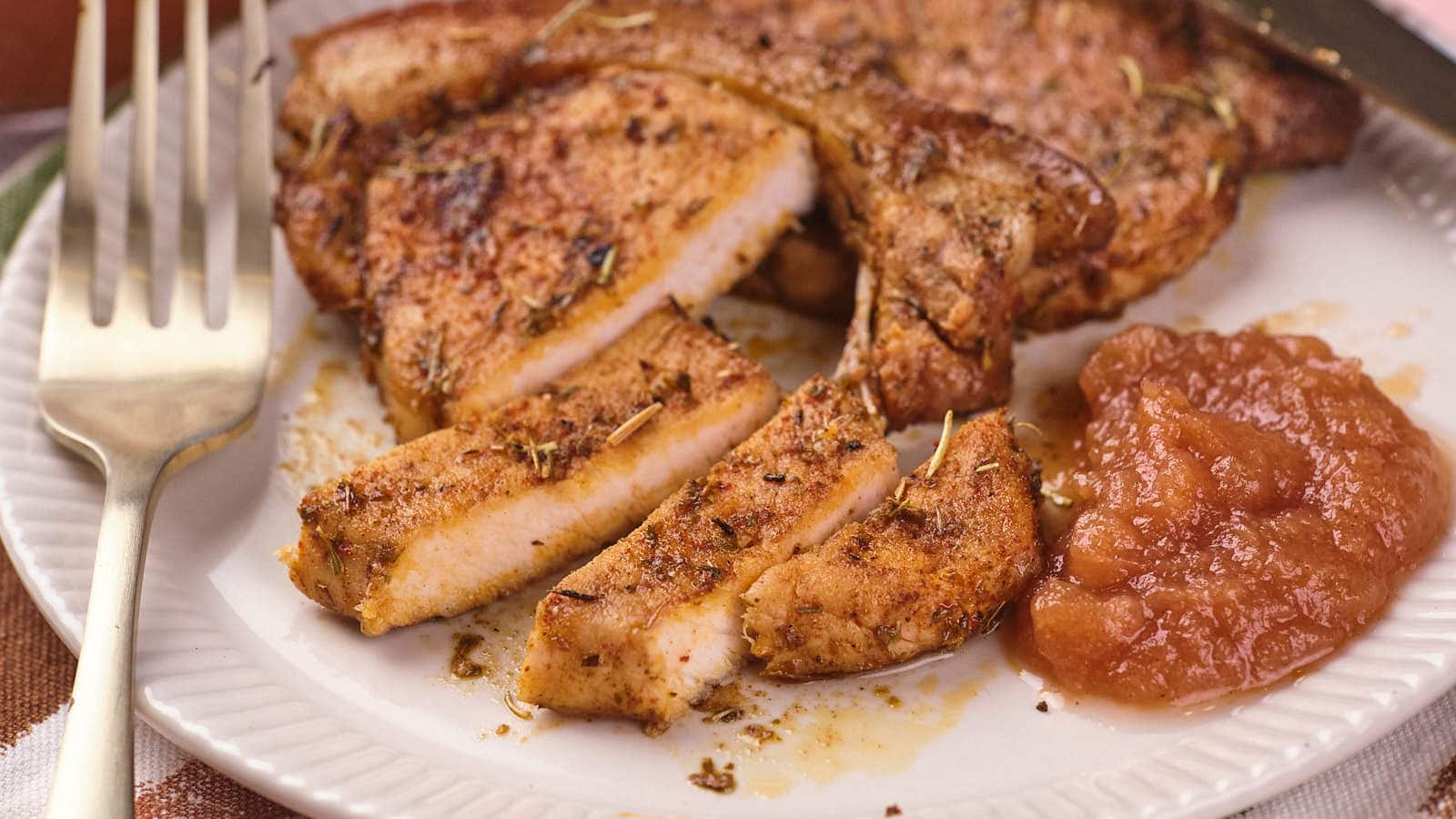 Seasoned pork chops sliced on a white plate with a serving of applesauce beside it and a fork nearby.