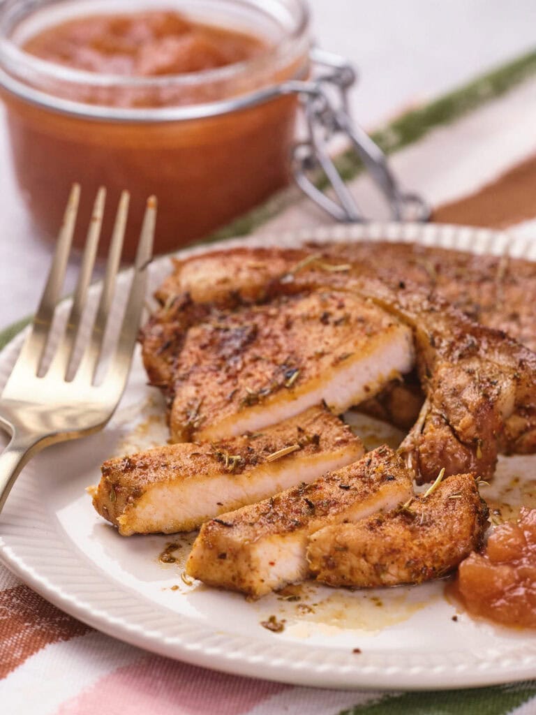 Sliced, seasoned pork chop on a white plate with a side of brown sauce and a gold fork.