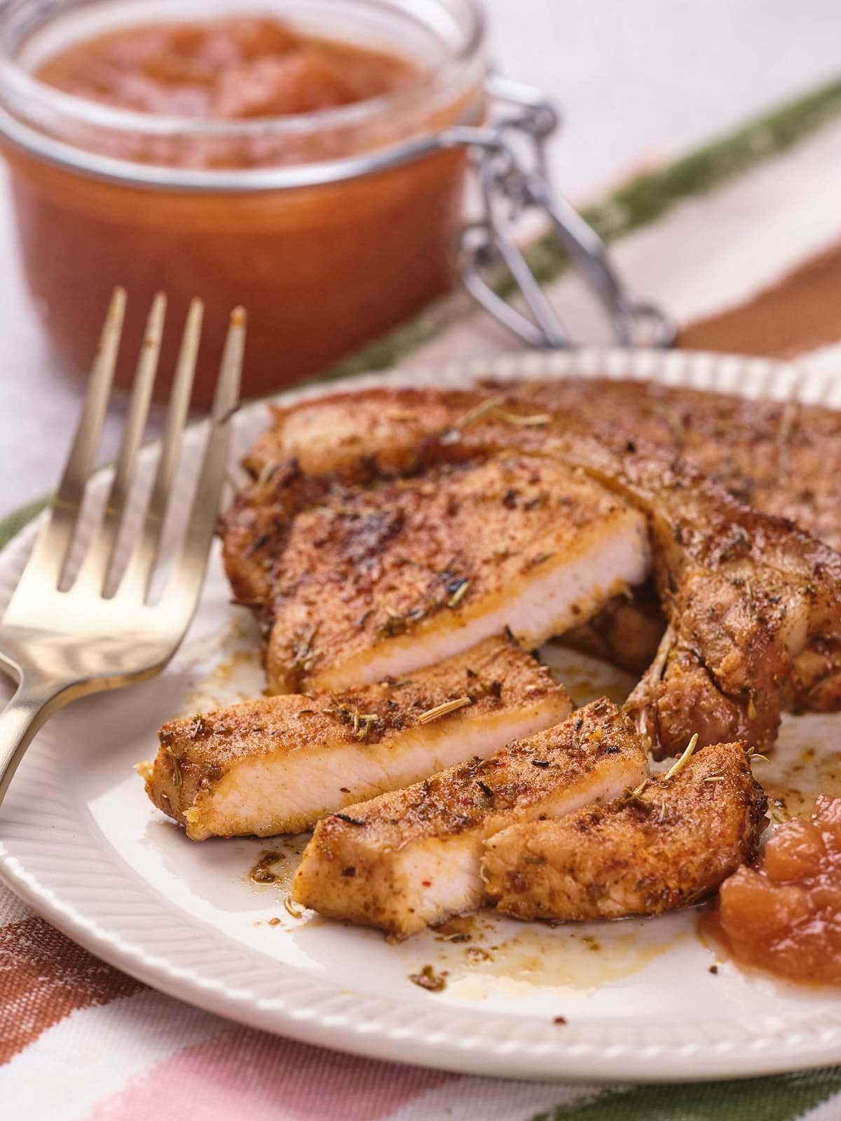 Sliced, seasoned pork chop on a white plate with a side of brown sauce and a gold fork.