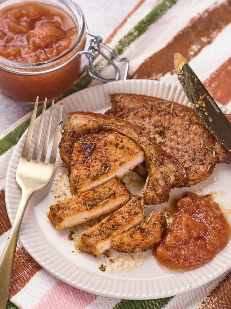 A plate with seasoned, sliced pork chop, a dollop of applesauce, a fork and knife, and a jar of applesauce in the background.