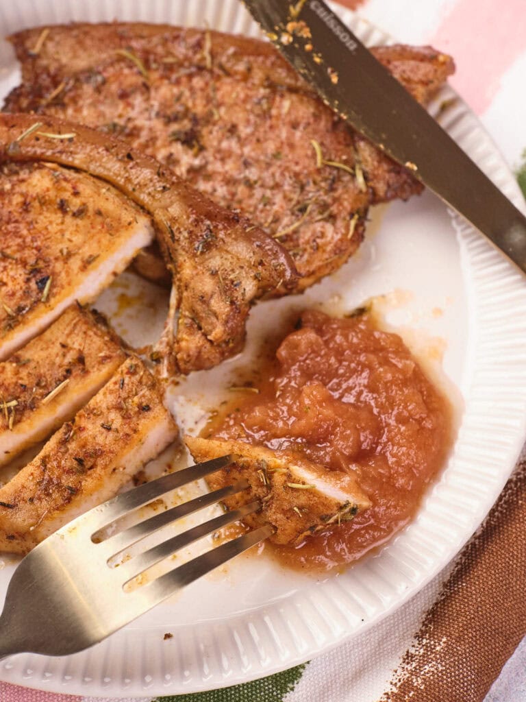 A cooked pork chop partially sliced on a white plate, served with a portion of applesauce.