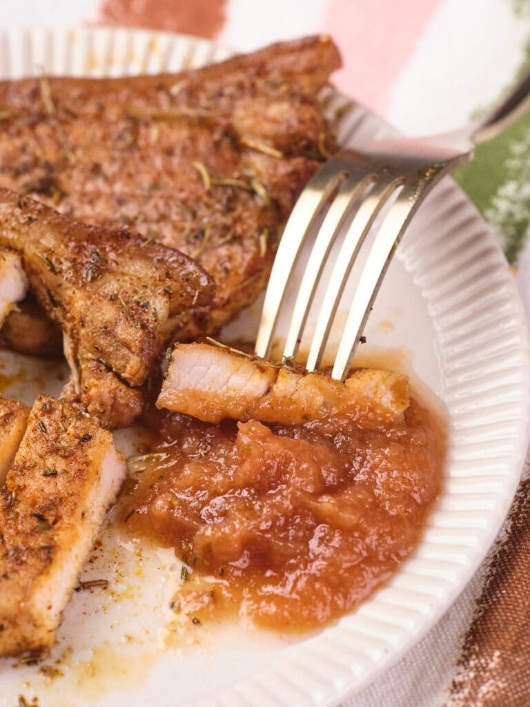 A fork holds a piece of seasoned pork chop dipped in applesauce on a white plate.