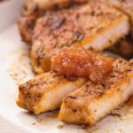Close-up of seasoned, cooked pork chops on a plate, with two slices topped with a dollop of apple sauce.
