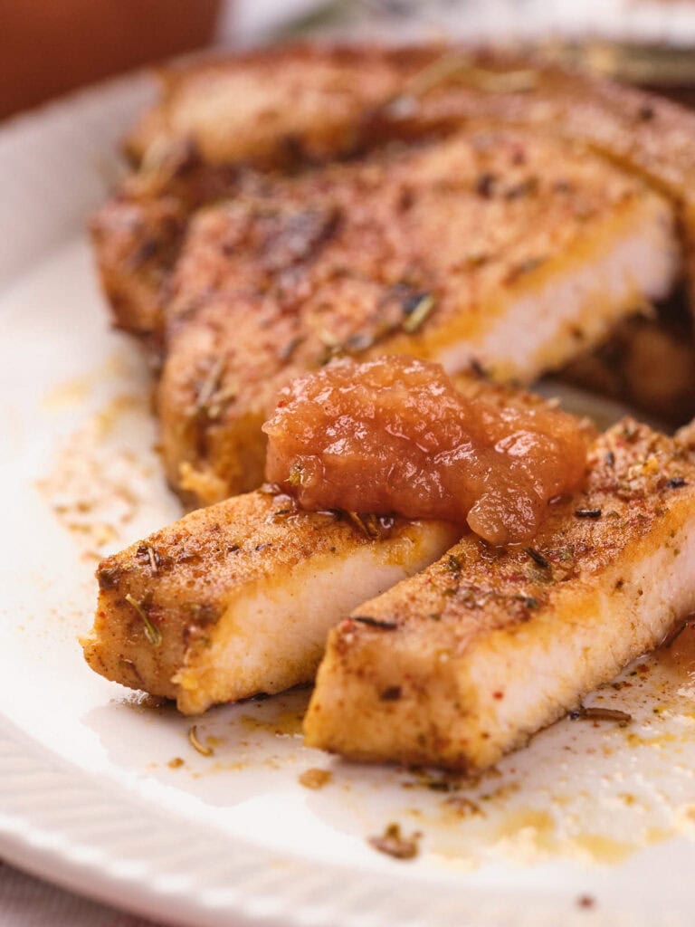 Close-up of seasoned, cooked pork chops on a plate, with two slices topped with a dollop of apple sauce.