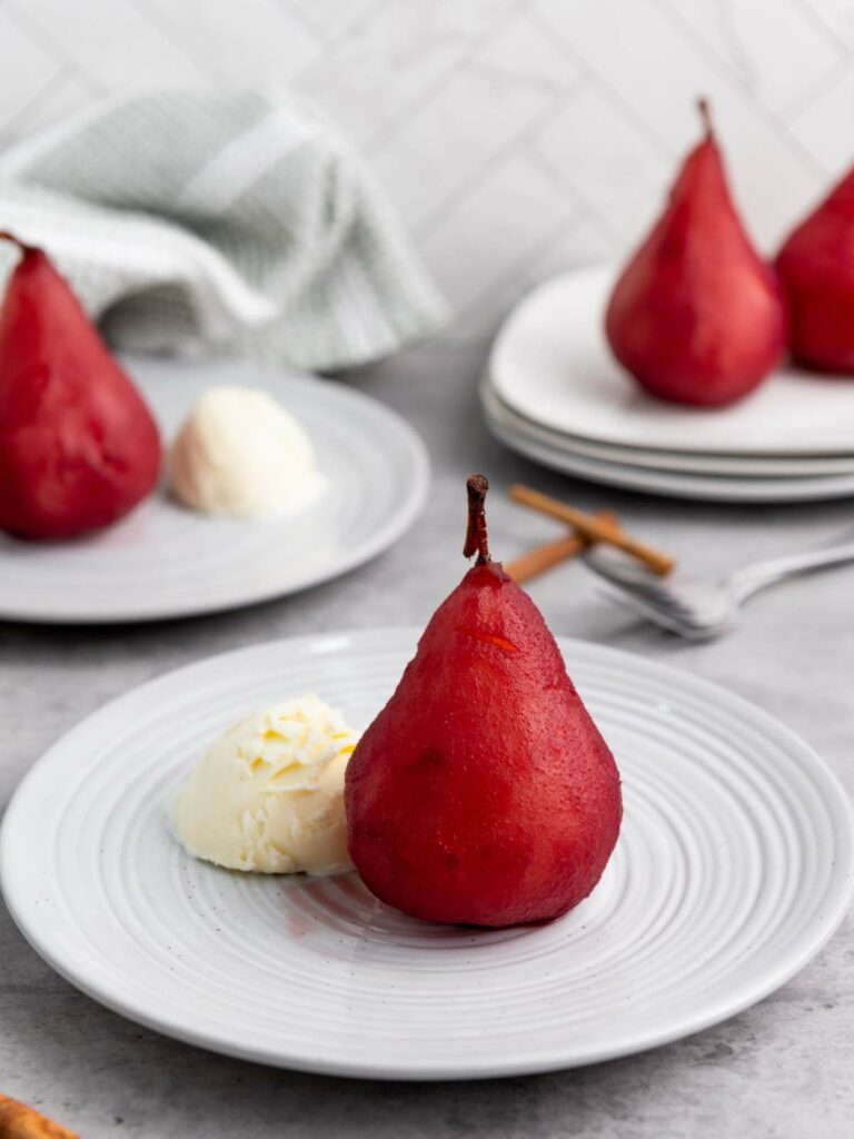 Poached pear and a scoop of vanilla ice cream are served on a white plate, with more pears and plates in the background.