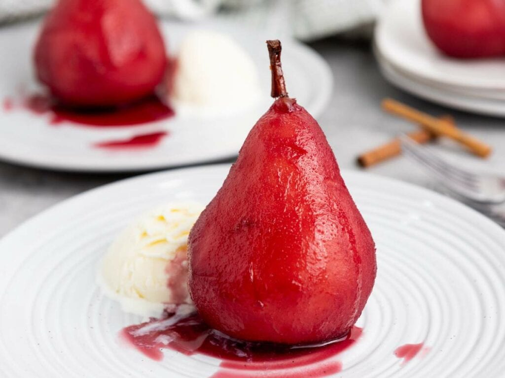 A pear in red sauce is plated with a scoop of vanilla ice cream on a white dish.