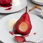 A pear with red sauce is served on a white plate with a scoop of vanilla ice cream.