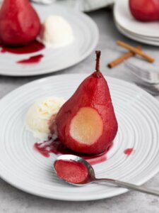 A pear with red sauce is served on a white plate with a scoop of vanilla ice cream.