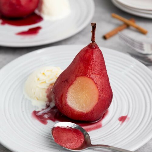 A pear with red sauce is served on a white plate with a scoop of vanilla ice cream.