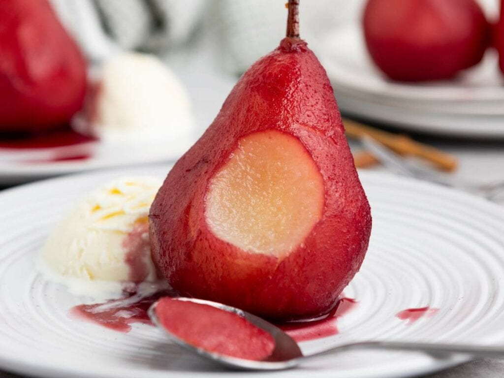A red pear with a bite taken out, served with a scoop of vanilla ice cream and sauce, on a white plate with a spoon in the foreground.