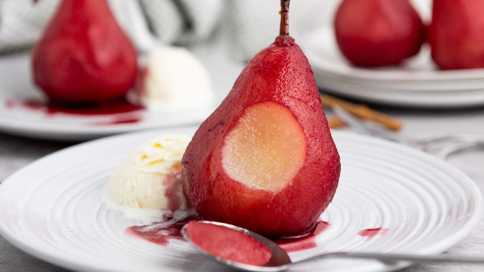 A poached pear with a missing bite sits on a white plate next to a scoop of vanilla ice cream, with a spoon and additional pears in the background.