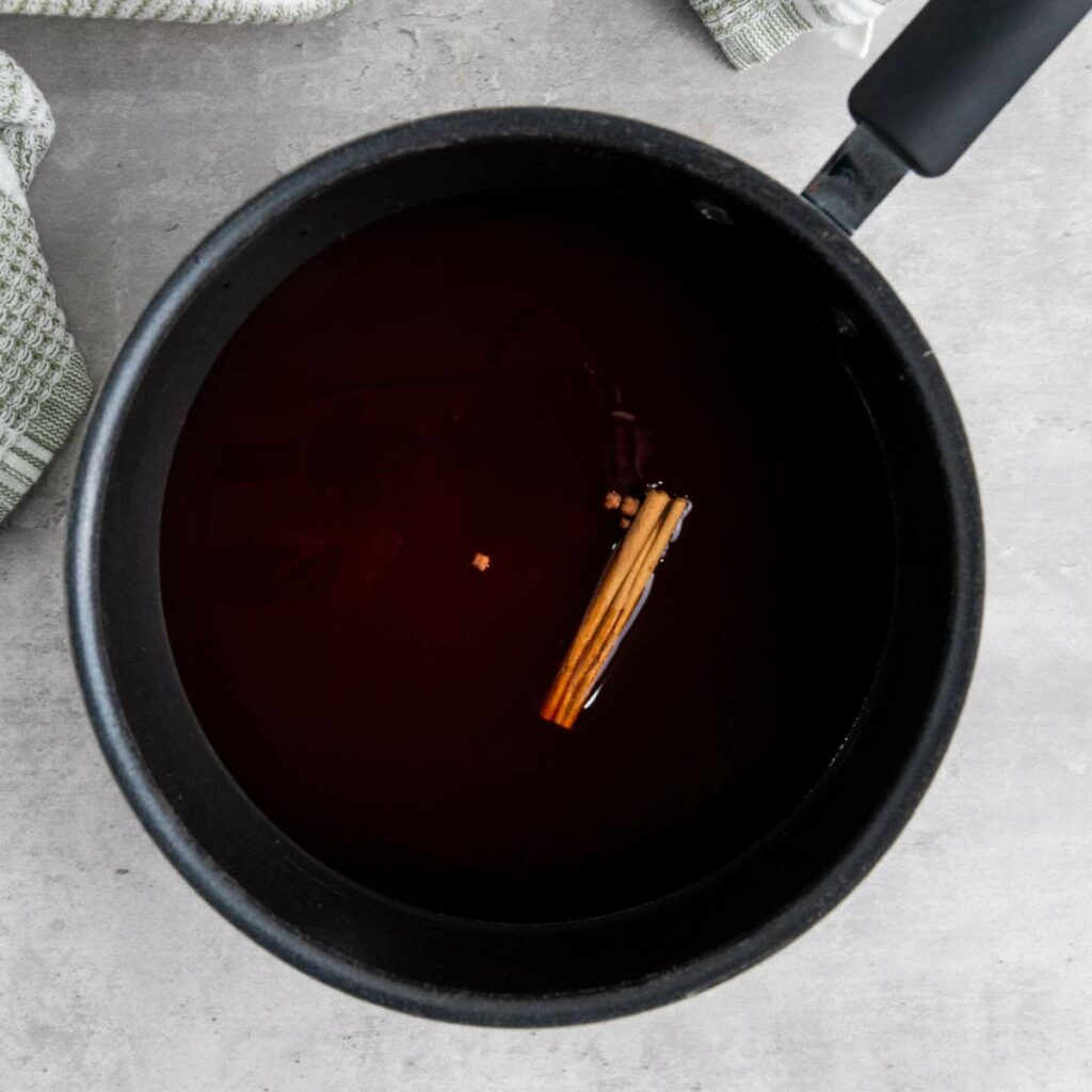 A saucepan filled with a dark liquid and a cinnamon stick floating in it, placed on a light gray surface.