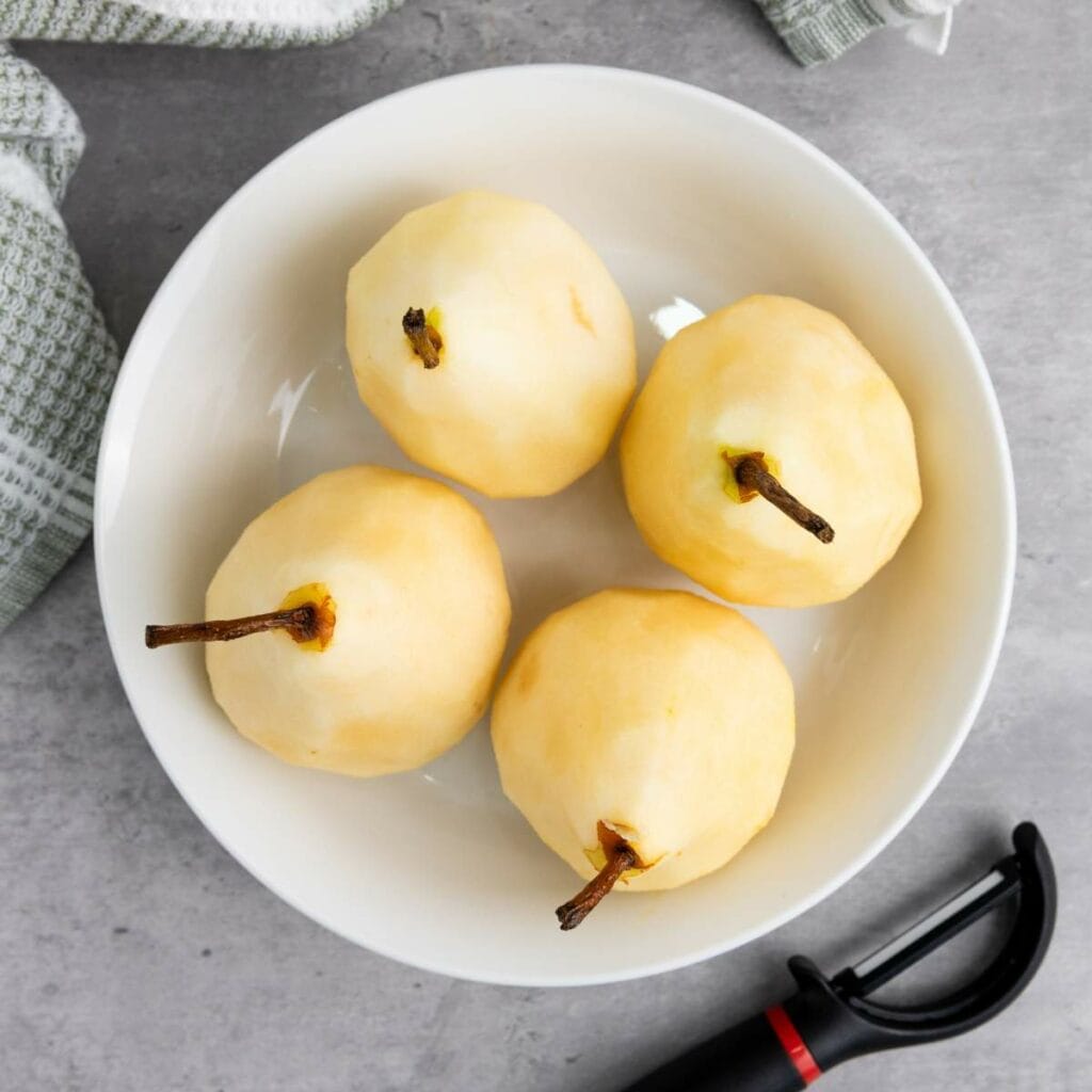 A white bowl containing four peeled pears with stems on a gray surface, next to a vegetable peeler and a textured cloth.