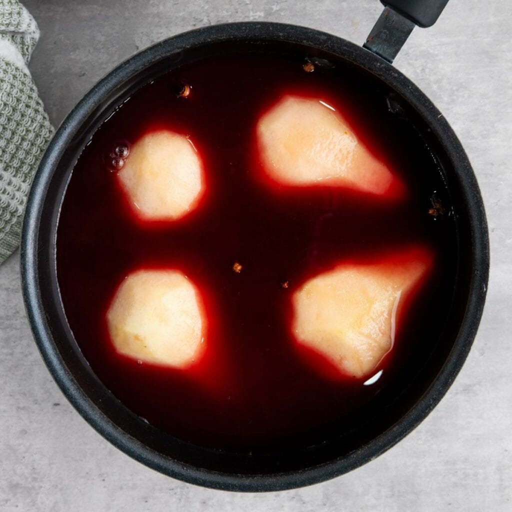 Four peeled pears are poaching in a pot of red wine on a light gray surface.