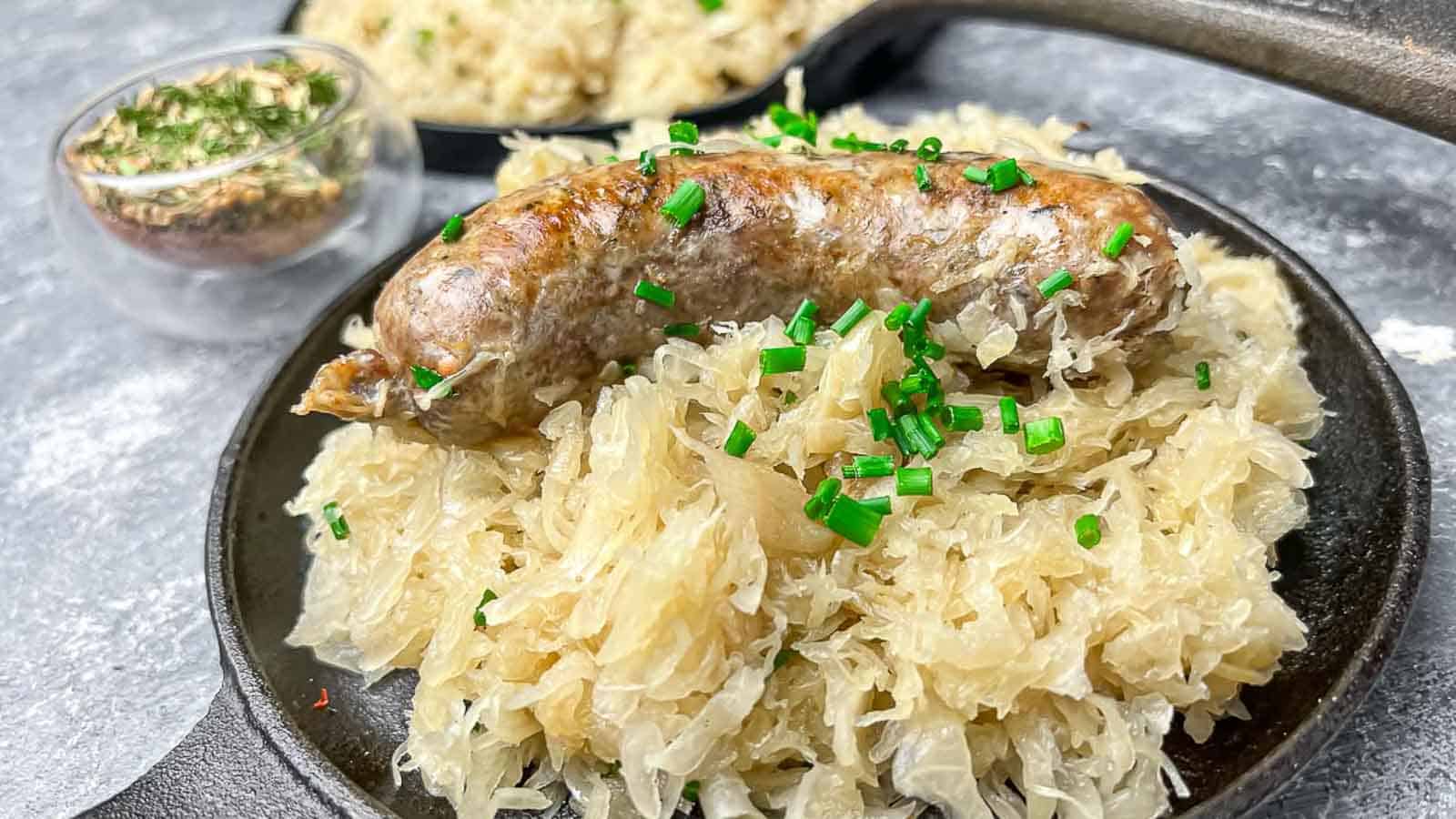 A cooked sausage served on a bed of sauerkraut, garnished with chopped chives, on a black plate. A small bowl of herbs and another plate of sauerkraut are in the background.
