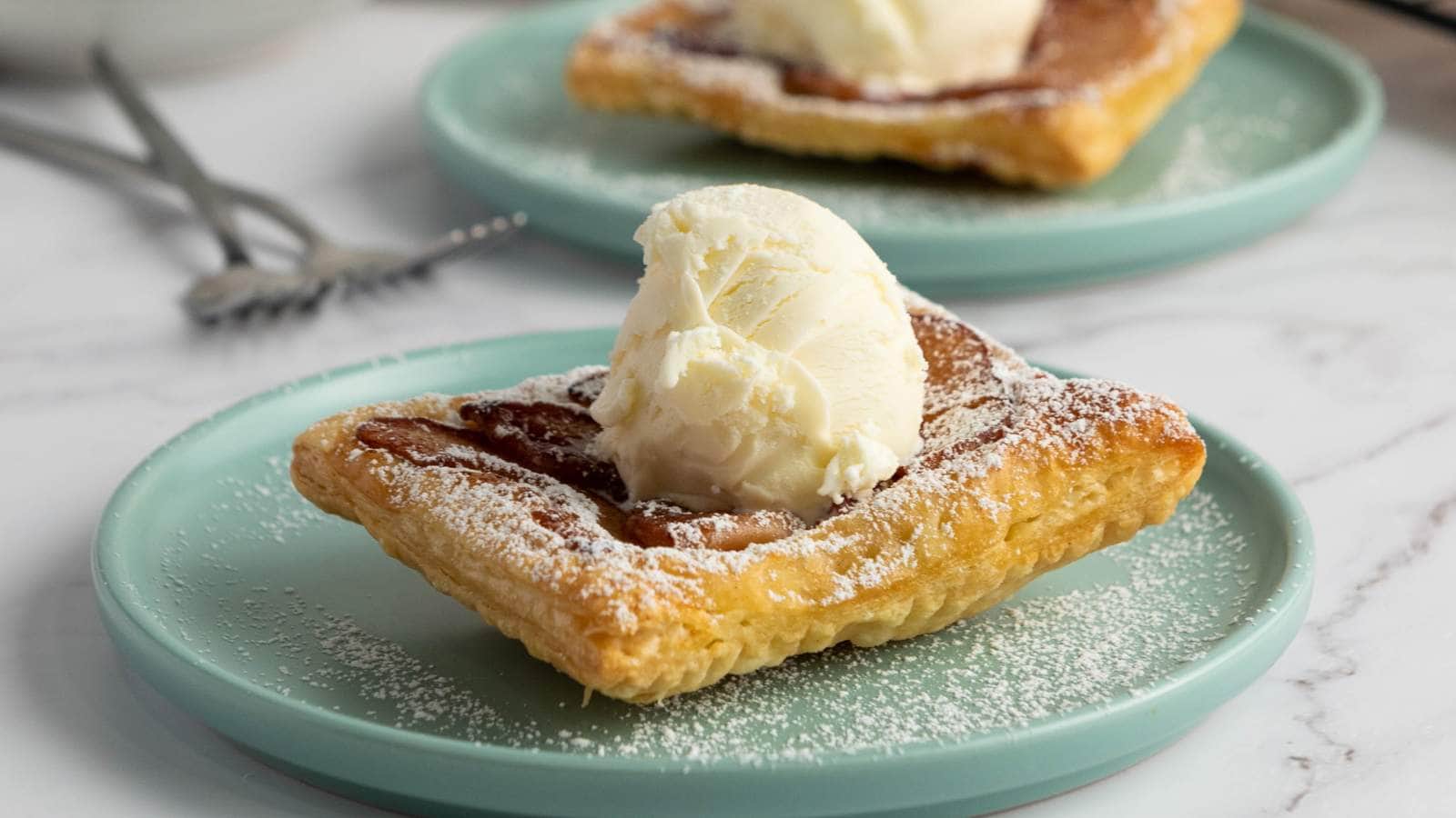 An aplpe tart with a scoop of vanilla ice cream and dusted with powdered sugar on a light blue plate.