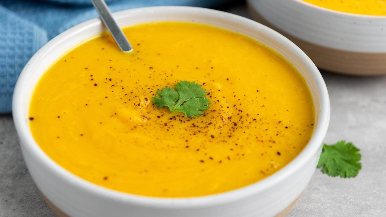 A bowl of smooth butternut squash soup, garnished with black pepper and a sprig of cilantro, with a spoon resting inside the bowl.