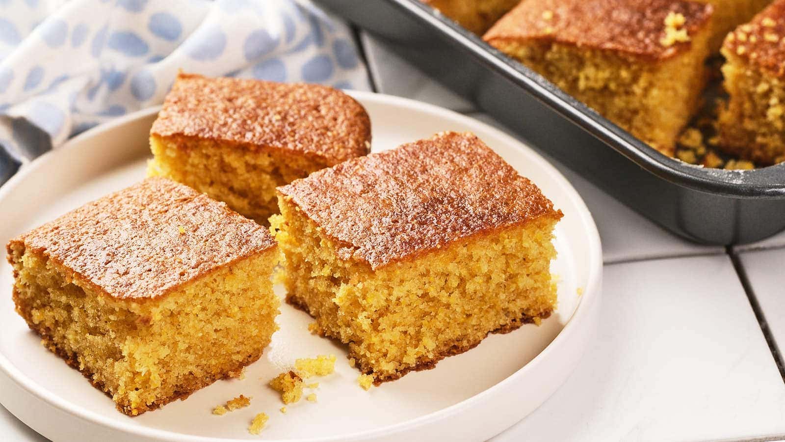 Three square pieces of cornbread are on a white plate, with more pieces visible in a baking pan in the background.