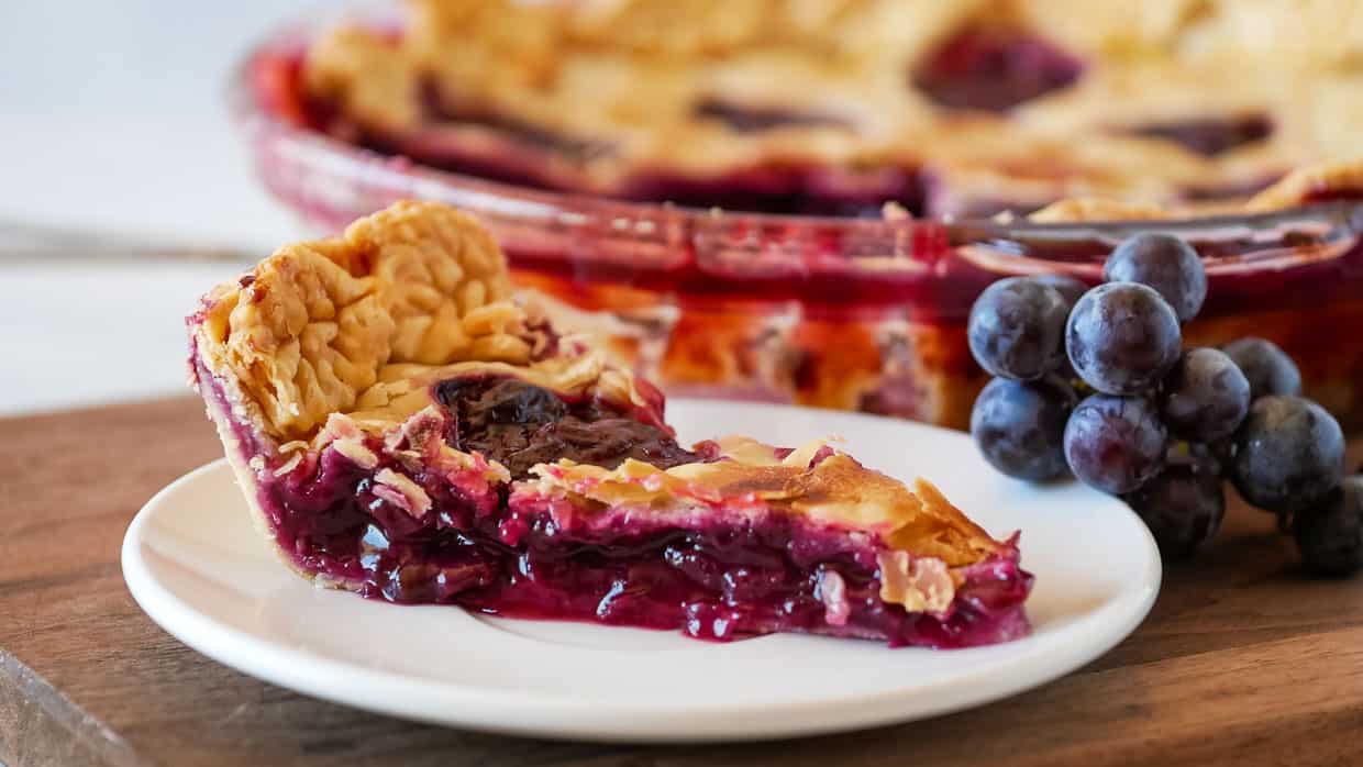 A slice of grape pie with a golden crust sits on a white plate, with a cluster of fresh grapes beside it and the rest of the pie in the background.