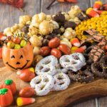 A wooden board with assorted Halloween treats, including candy corn, popcorn, chocolate-covered pretzels, and festive candies, with a pumpkin container and autumn leaves.