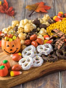 A wooden board with assorted Halloween treats, including candy corn, popcorn, chocolate-covered pretzels, and festive candies, with a pumpkin container and autumn leaves.