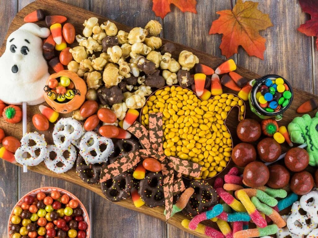 A wooden board filled with assorted Halloween-themed candies, pretzels, popcorn, and treats, surrounded by autumn leaves on a wooden surface.