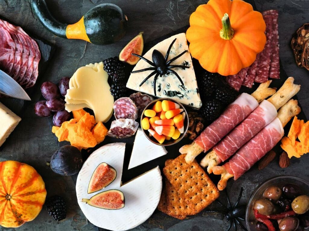 A Halloween-themed charcuterie board with assorted cheeses, meats, crackers, figs, grapes, pumpkins, candy corn, and a fake spider on top of blue cheese.