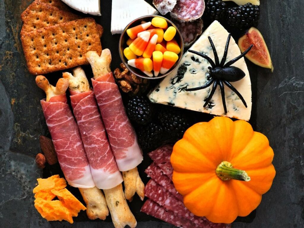 A Halloween-themed charcuterie board with crackers, prosciutto-wrapped breadsticks, cheese, candy corn, blackberries, salami, a pumpkin, and a plastic spider.