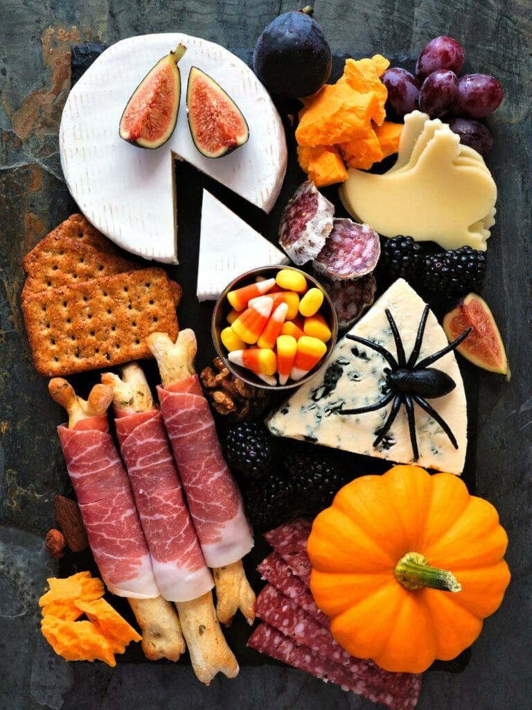 A Halloween-themed charcuterie board with assorted cheeses, crackers, meats, grapes, figs, candy corn, a small pumpkin, and a plastic spider.