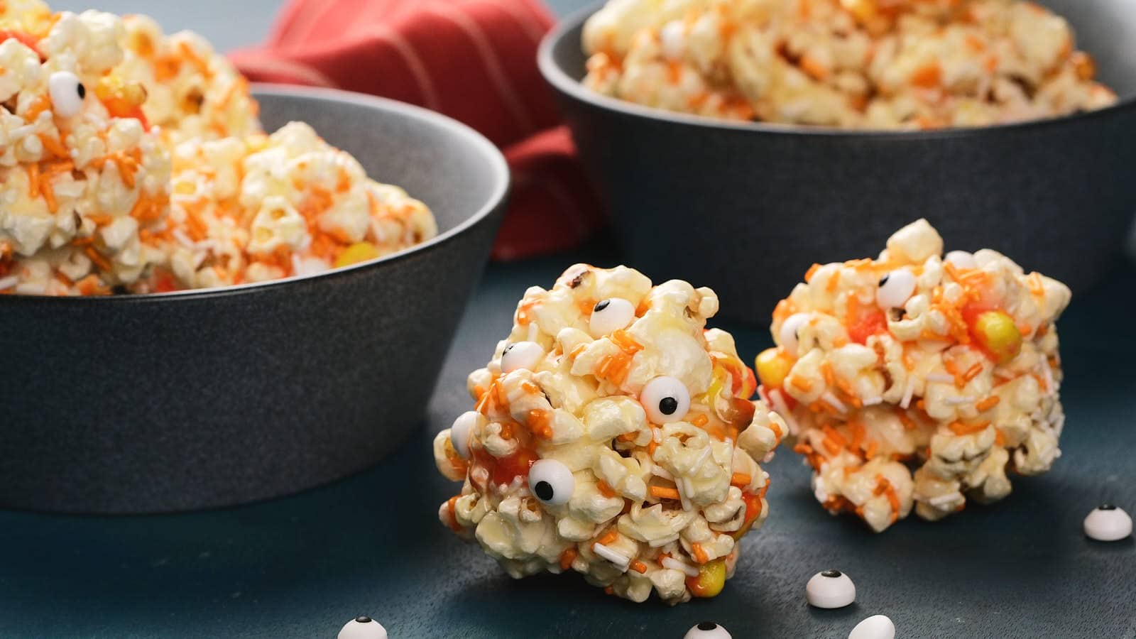 Popcorn balls decorated with candy eyes, candy corn and orange sprinkles sit on a table next to bowls filled with more popcorn balls.