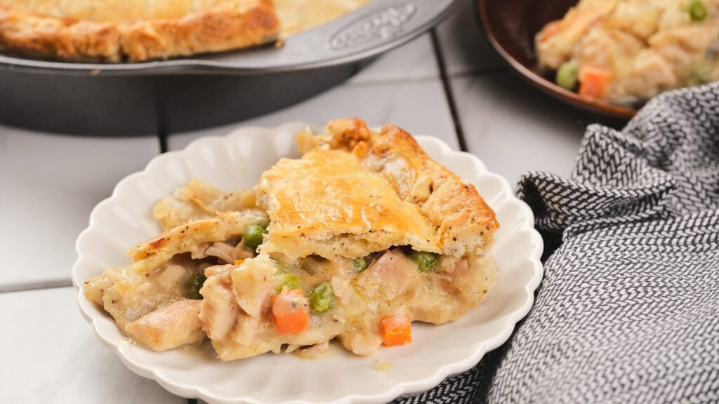A serving of chicken pie with a golden crust on a white plate, showing pieces of chicken, peas, and carrots, with more pie in the background.