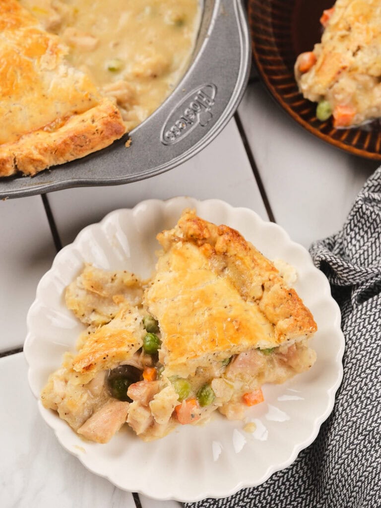 A slice of chicken pie served on a white plate, next to the remaining pie in a baking dish and a brown plate.