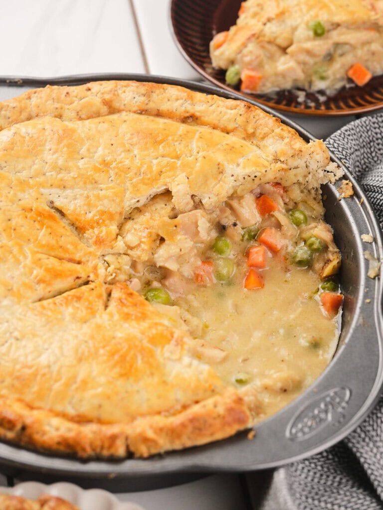 A baked chicken pot pie in a metal pie dish with a slice removed, showing a creamy filling with peas and carrots.