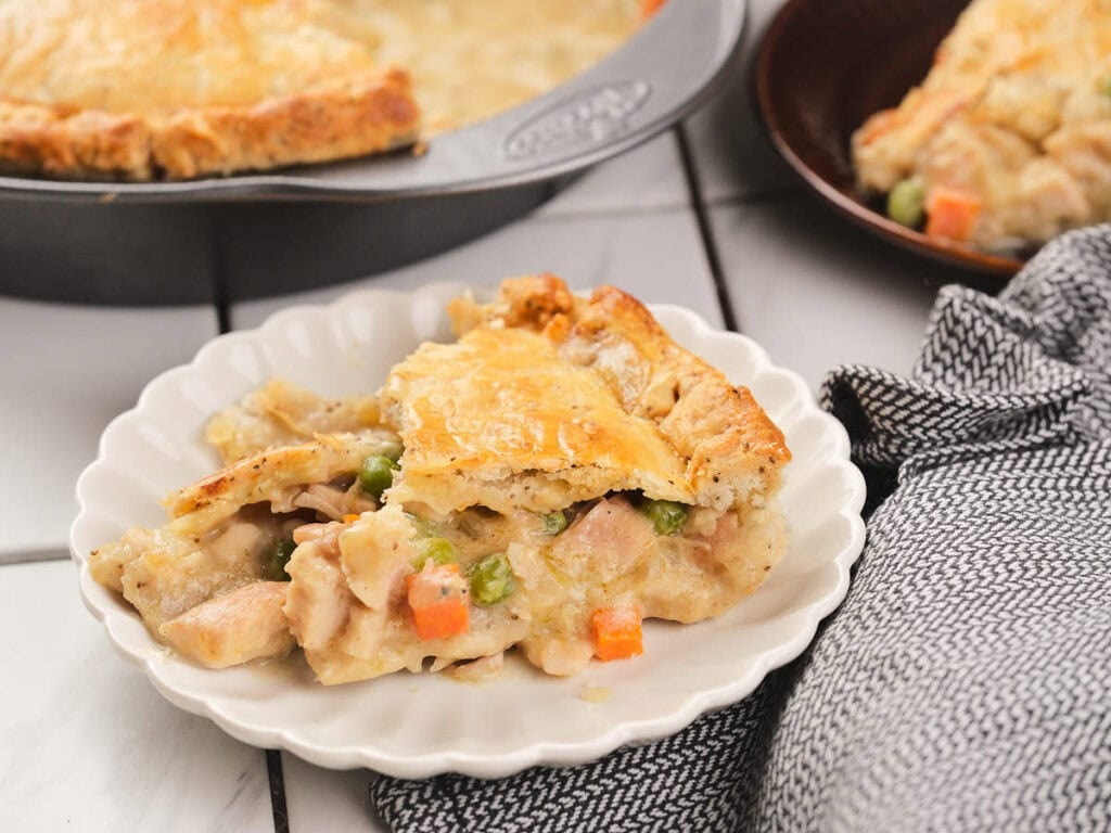 A slice of chicken pie with a golden crust on a white plate, showing chunks of chicken, peas, and carrots.