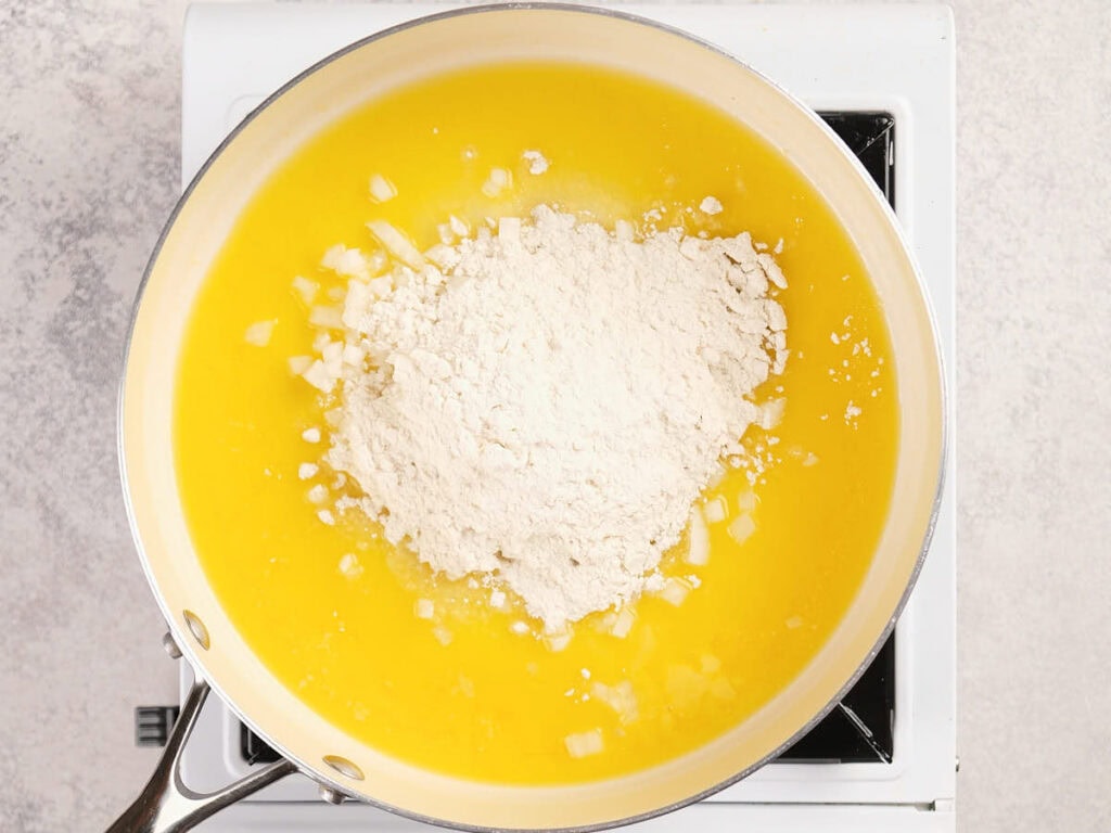 A saucepan on a stove contains melted butter, chopped onions, and a pile of flour, ready to be mixed.