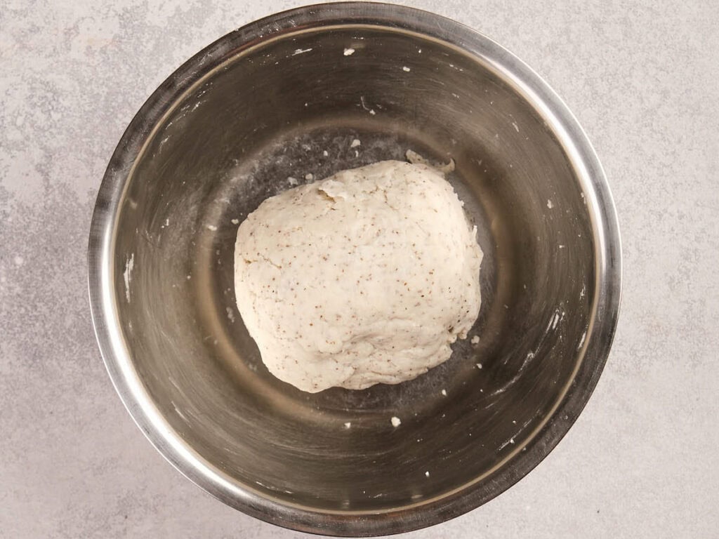 A ball of dough sits in a stainless steel mixing bowl on a light gray surface.
