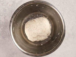 A ball of dough sits in a stainless steel mixing bowl on a light gray surface.